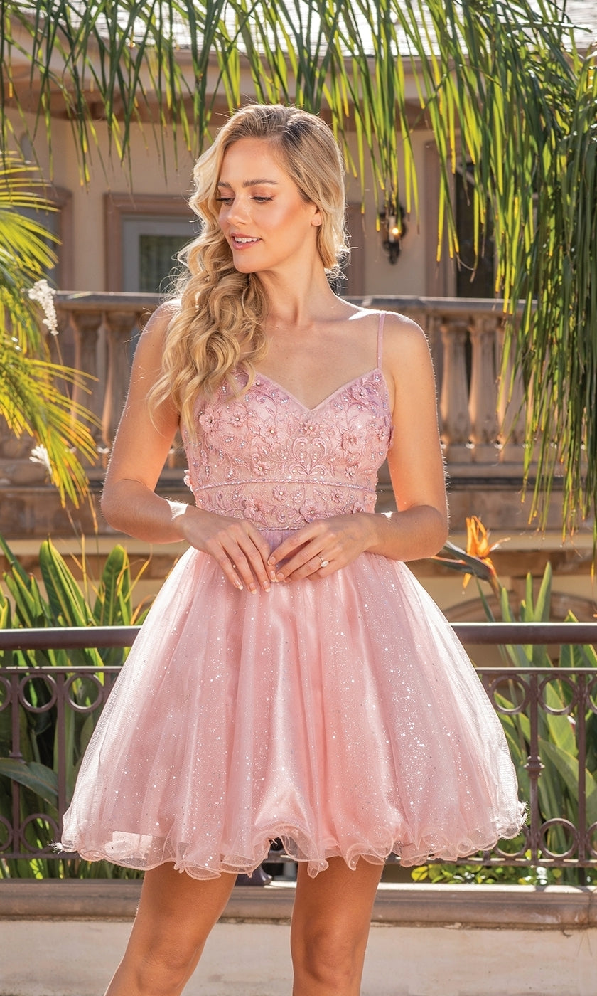 A woman stands outdoors in the Glitter-Tulle Short A-Line Homecoming Dress 3336, a sleeveless light pink babydoll style with an embellished V-neck bodice and tulle skirt. Green palm leaves and a building are in the background.
